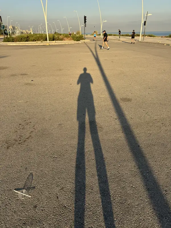A runner's long shadow stretched across an empty seafront road in the sun.